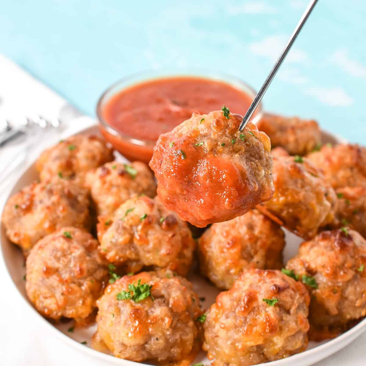 A close up image of a ball held up by a metal toothpick and dipped in the marinara sauce. The plate with the rest and the sauce is in the background.