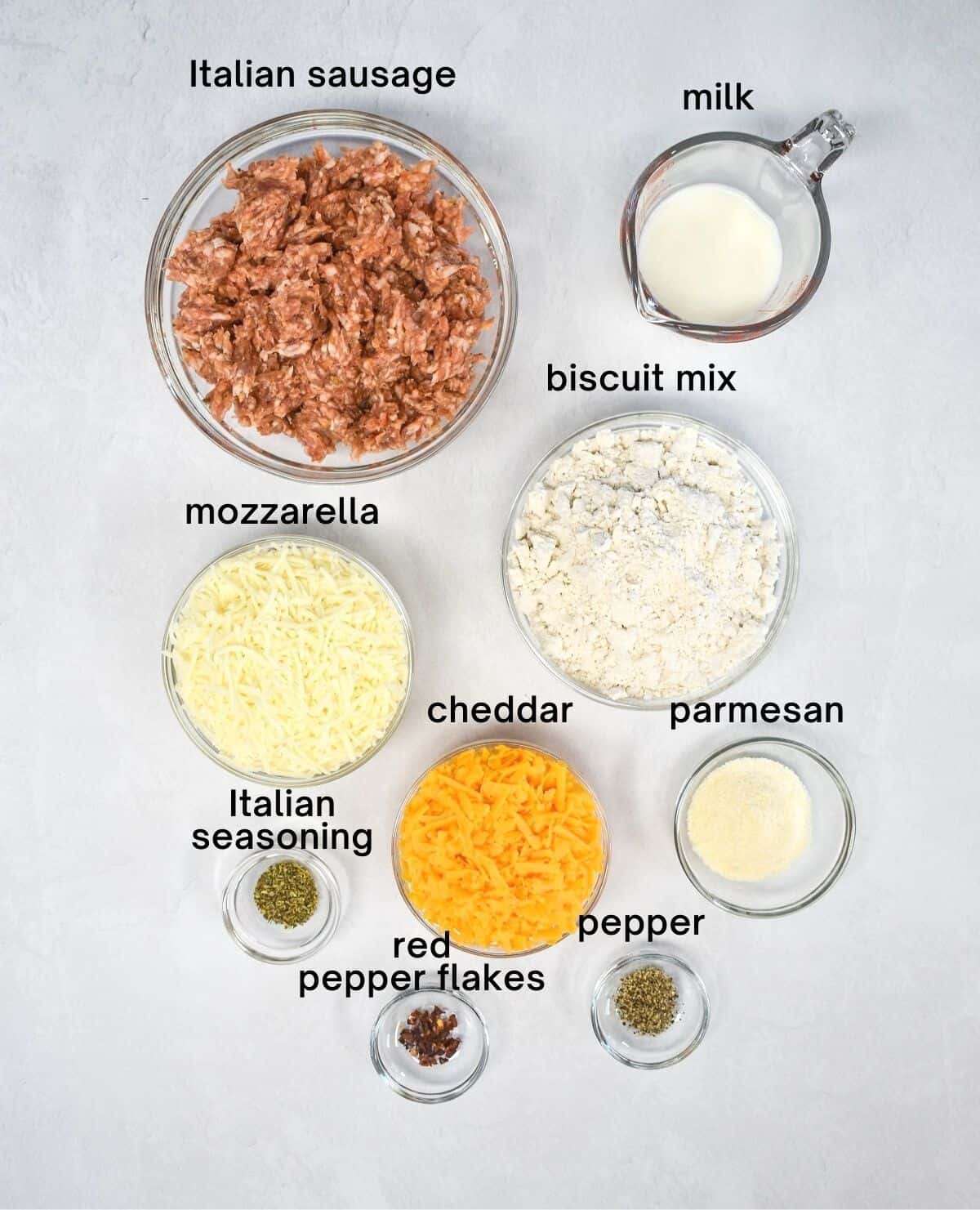 The ingredients for the sausage balls arranged in clear bowls and set on a white table. Each ingredient has a label above in black letters.