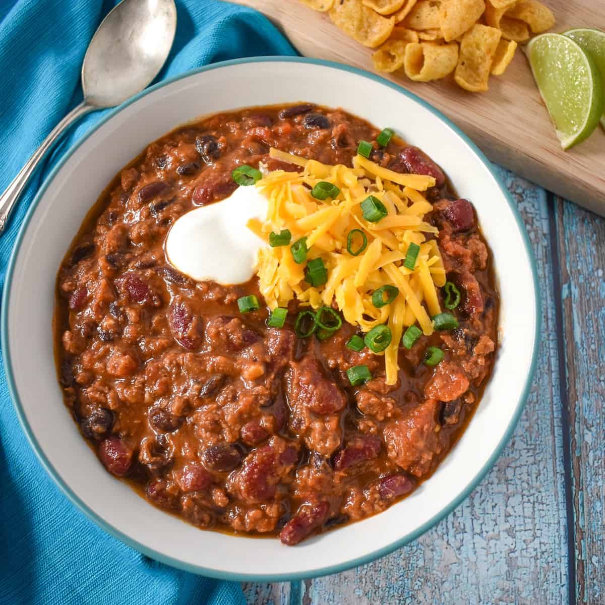 An image of the finished chili in a white bowl garnished with sour cream, cheese and green onions. The bowl is set with an aqua colored linen with corn chips and lime in the background.