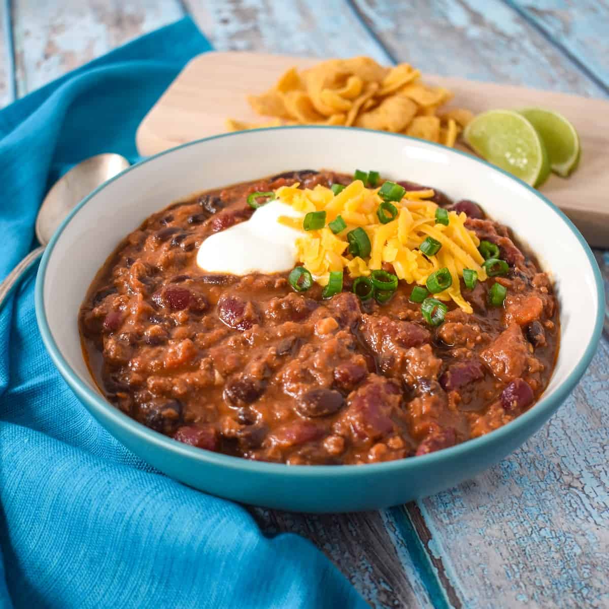 An image of the beef and bean chili in a white bowl garnished with sour cream, cheese and green onions. The bowl is set with an aqua colored linen with corn chips and lime in the background.