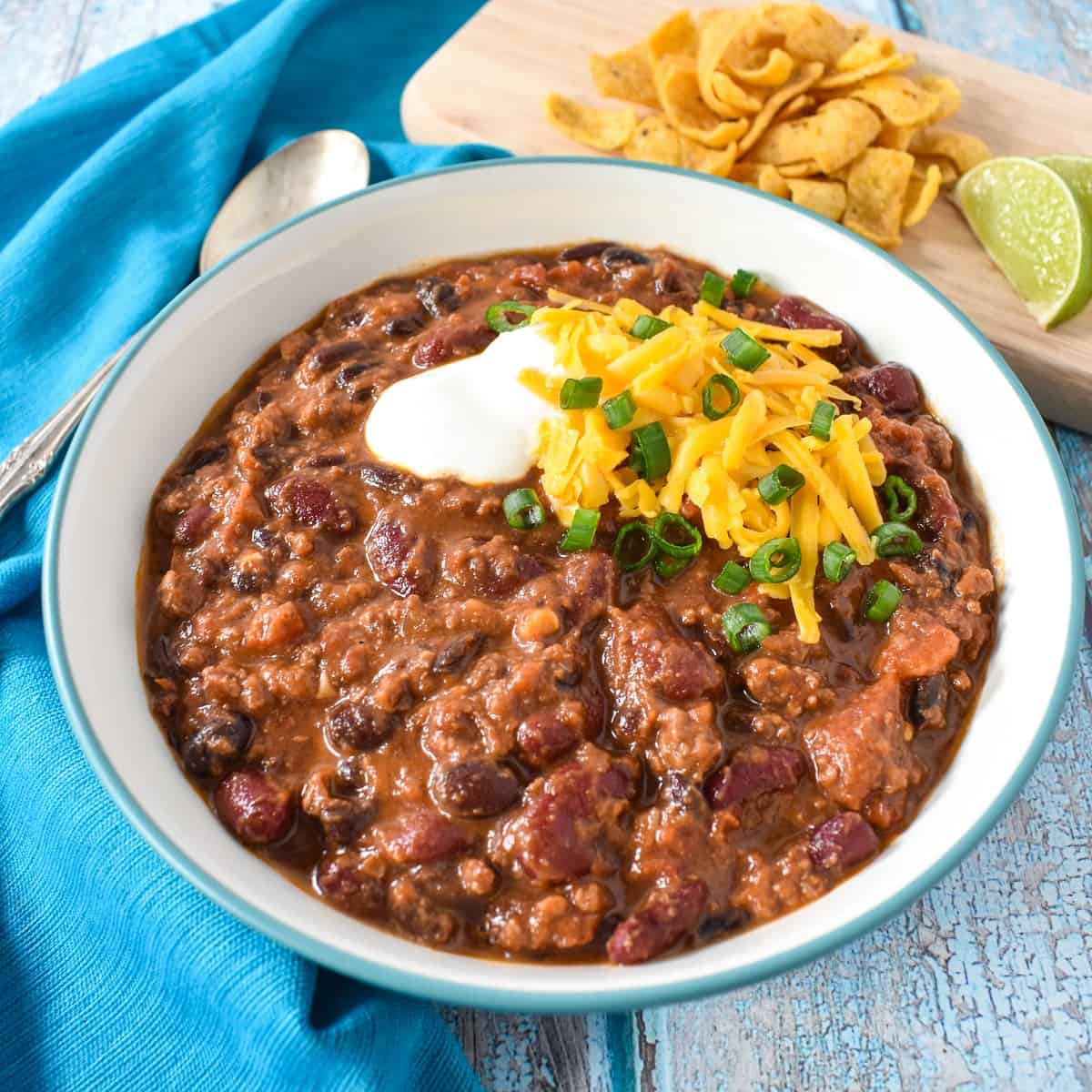 An image of the beef chili with beans served in a white bowl garnished with sour cream, cheese, and green onions. The bowl is set with an aqua colored linen with corn chips and lime in the background.