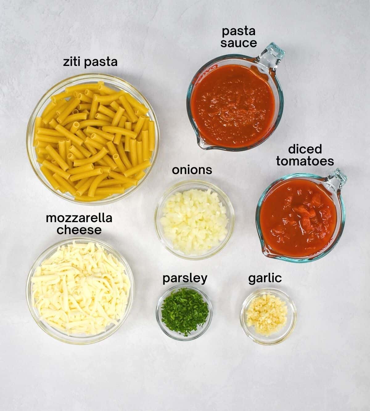 The ingredients for the sauce, pasta and casserole arranged in clear bowls on a white table.