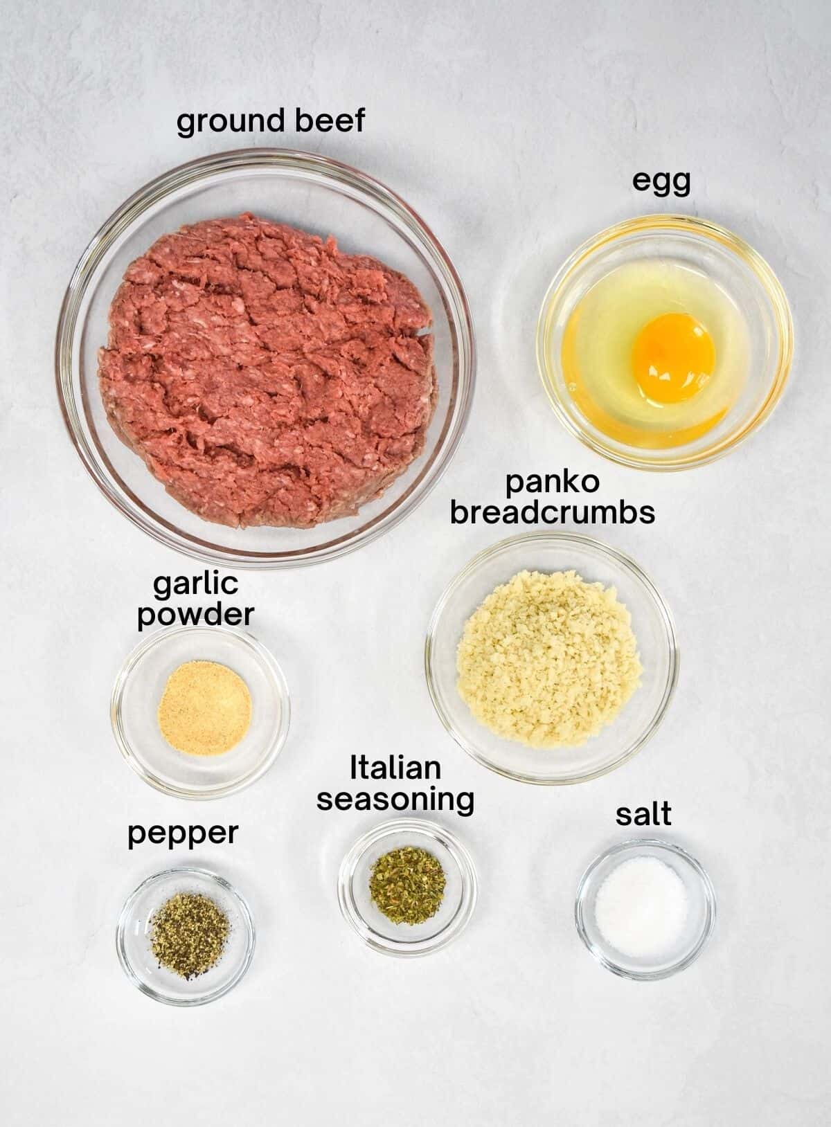 An image of the ingredients for the meatballs separated in clear glass bowls and set on a white table.