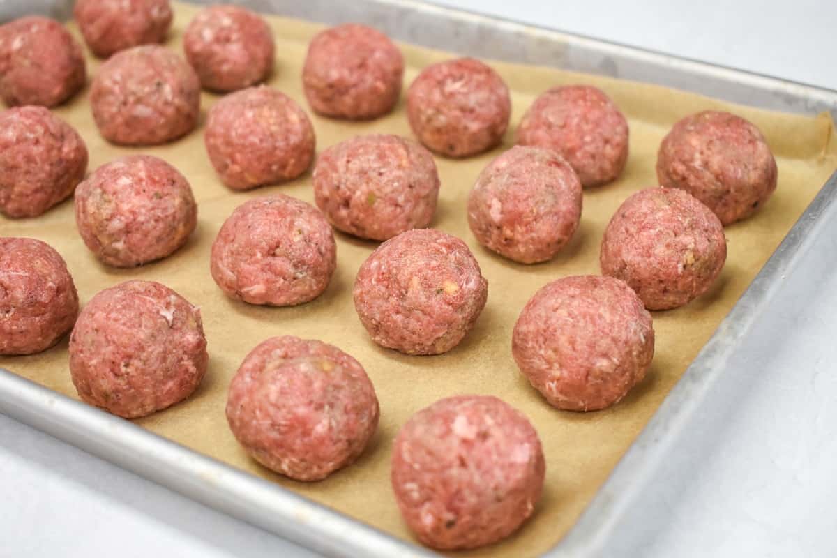 An image of meatballs, before cooking, arranged on a baking sheet lined with parchment paper.