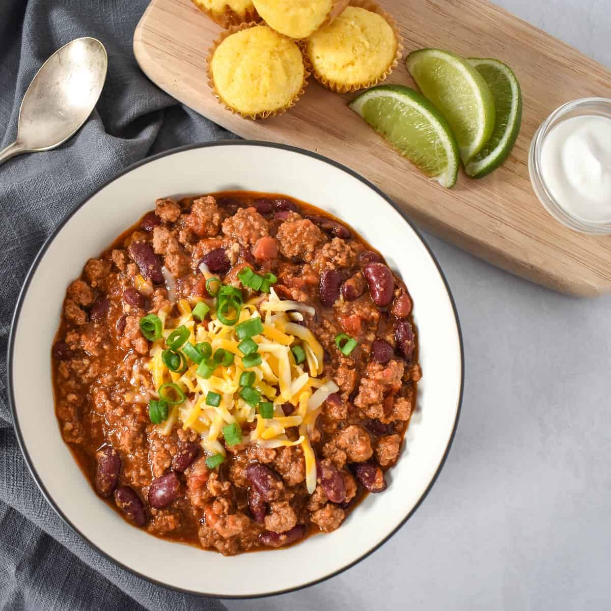 An image of the chili garnished with cheese and green onions and served with cornbread muffins, lime wedges, and sour cream on the side.