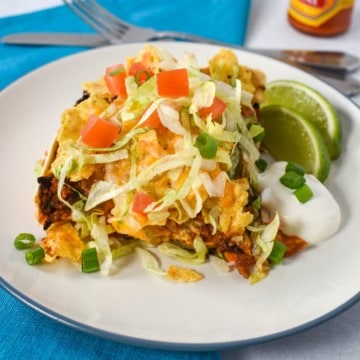 An image of the taco casserole with a side of lime wedges and sour cream served on a white plate set on an aqua linen.