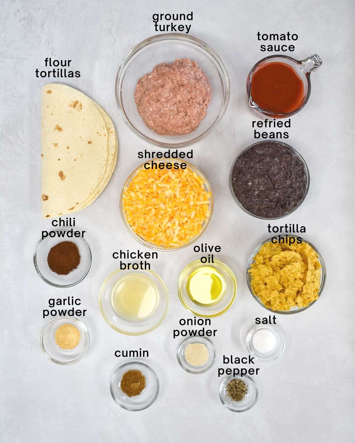 An image of the ingredients for the taco casserole arranged in glass bowls on a white table. Each ingredient is labled with small black letters.