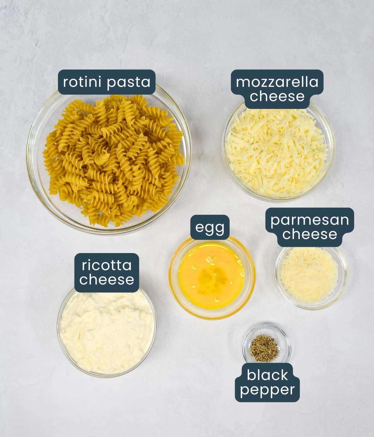 Ingredients for the pasta and cheese layers of easy lasagna casserole laid out and labeled: rotini pasta, mozzarella cheese, egg, Parmesan cheese, ricotta cheese, and black pepper.