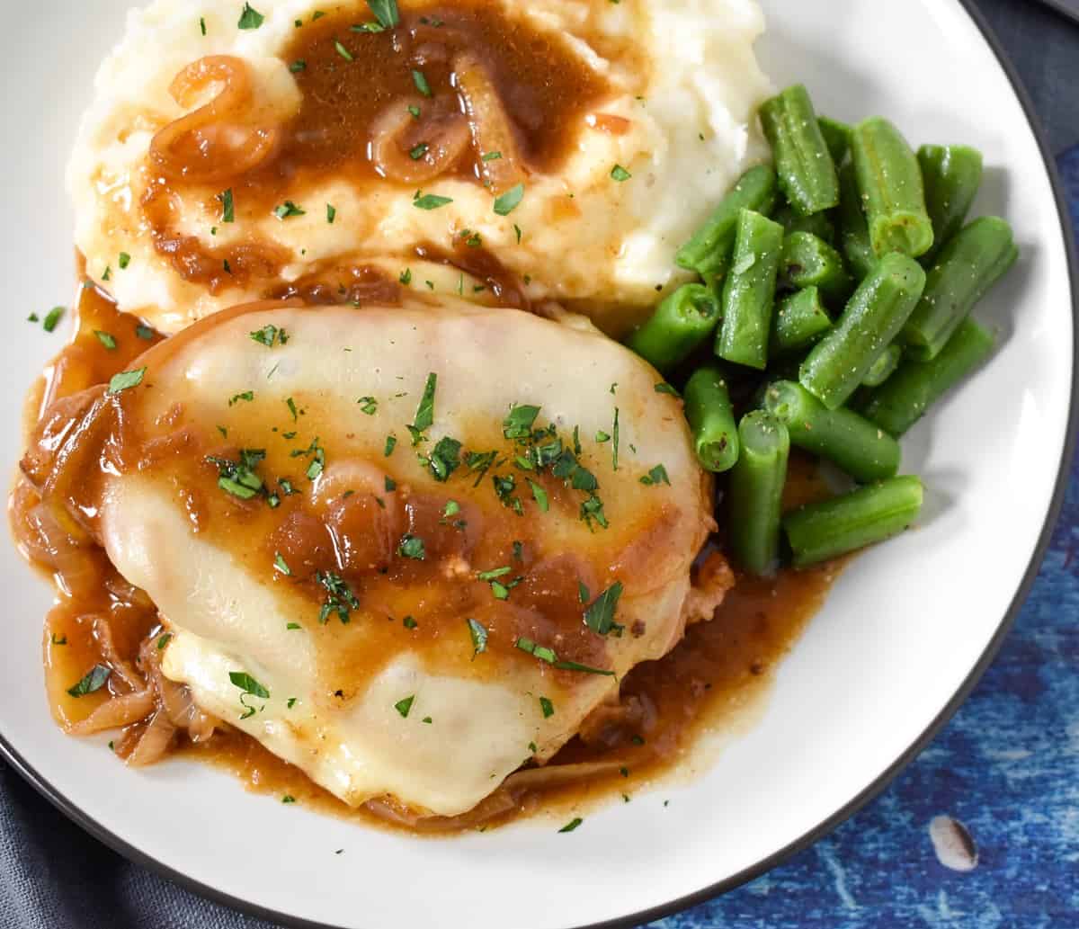 An image of the french onion pork chop served with mashed potatoes, covered with the onion sauce and a side of green beans.