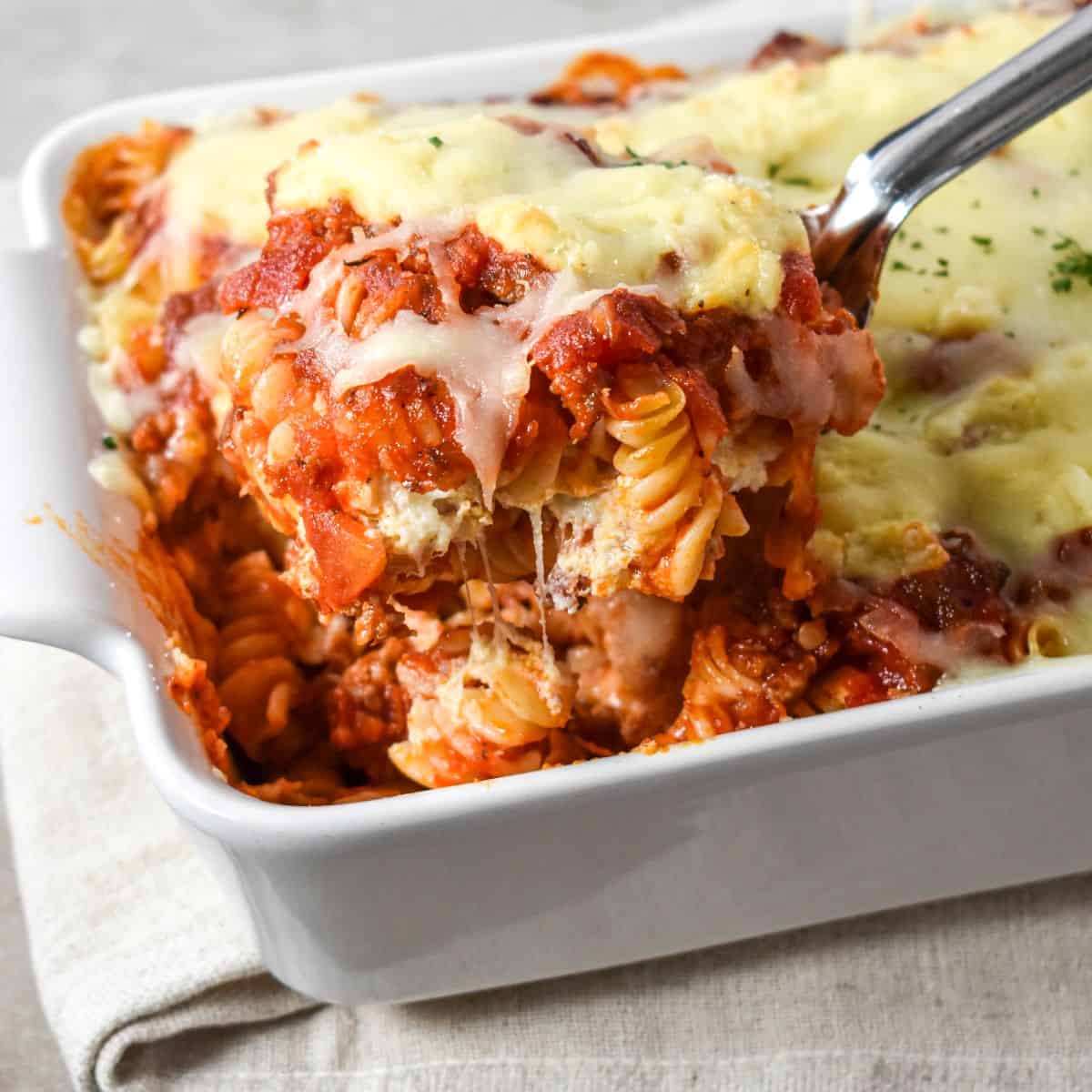 Easy lasagna casserole with layers of rotini, sausage sauce, ricotta, and melted mozzarella being served with a spoon from a white baking dish.