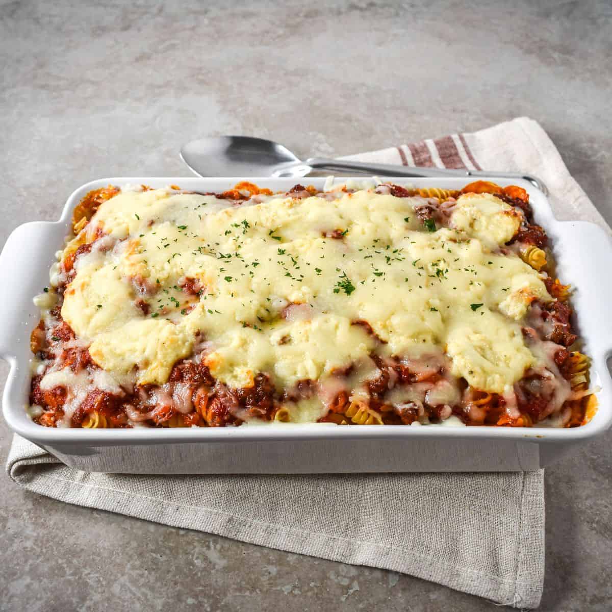 Easy lasagna casserole topped with melted mozzarella and chopped parsley in a white baking dish on a linen napkin, with a serving spoon behind it.