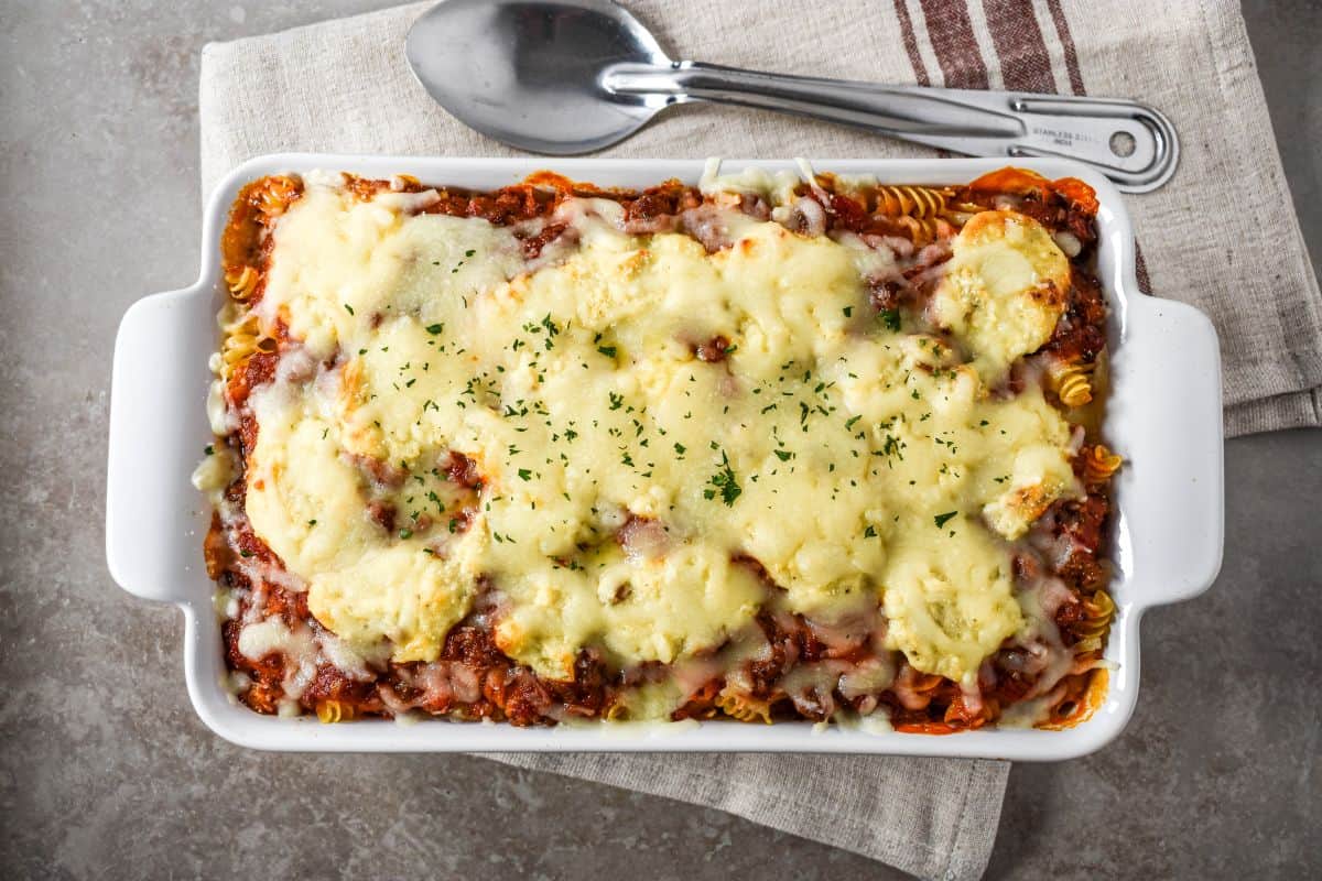 Baked easy lasagna casserole with melted mozzarella and parsley in a white rectangular baking dish, with a spoon on a linen napkin.