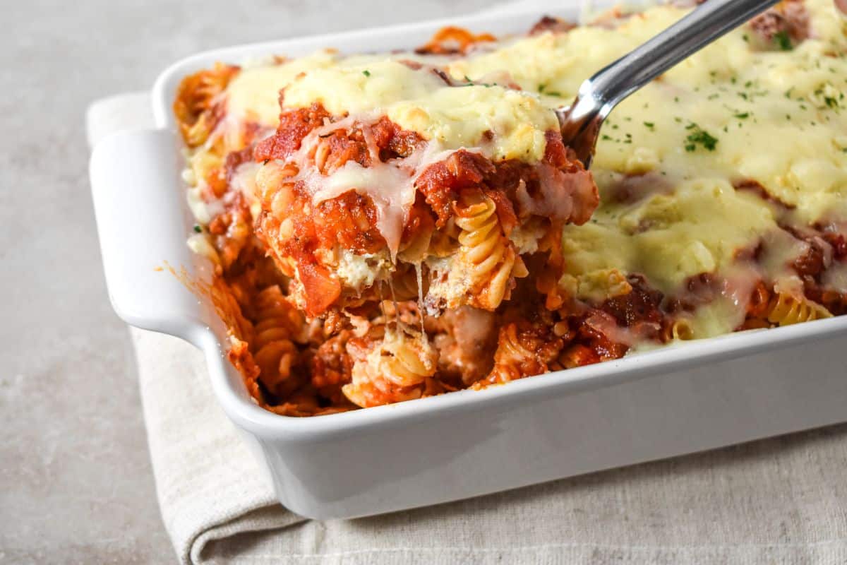 Easy lasagna casserole with layers of rotini, sausage sauce, ricotta, and melted mozzarella being served with a spoon from a white baking dish.