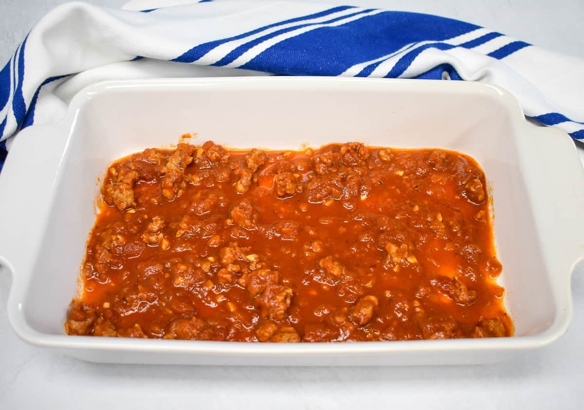 An image of a thin layer of meat sauce in a white baking dish with a blue and white dish towel.