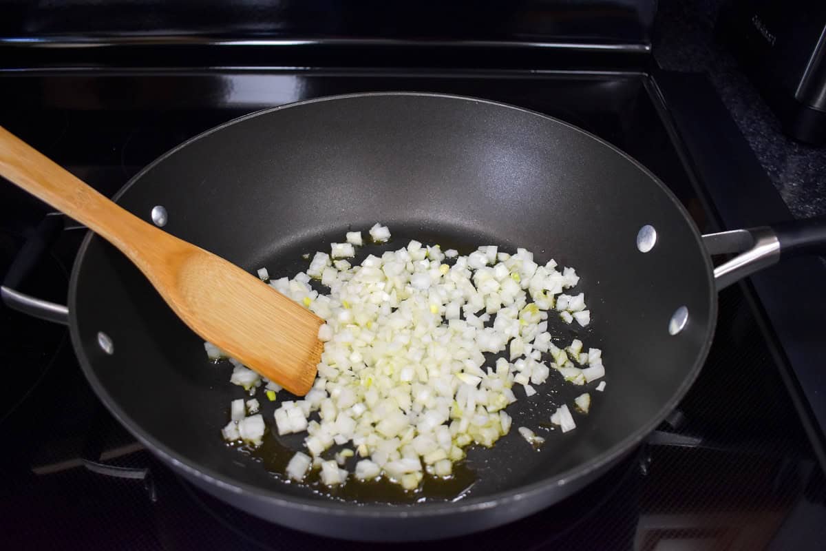 Diced onions cooking in olive oil in a large skillet with a wooden spoon.