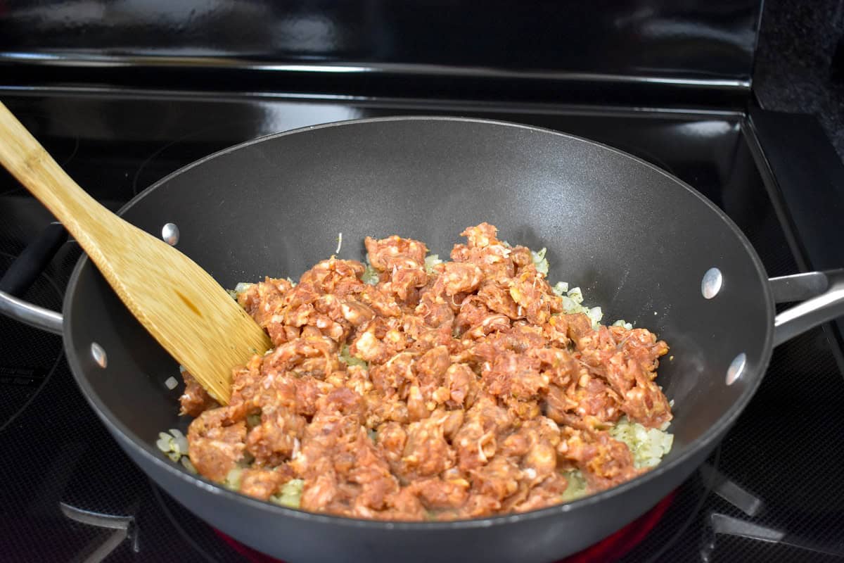 Raw Italian sausage added to the onions in a large skillet with a wooden spoon.