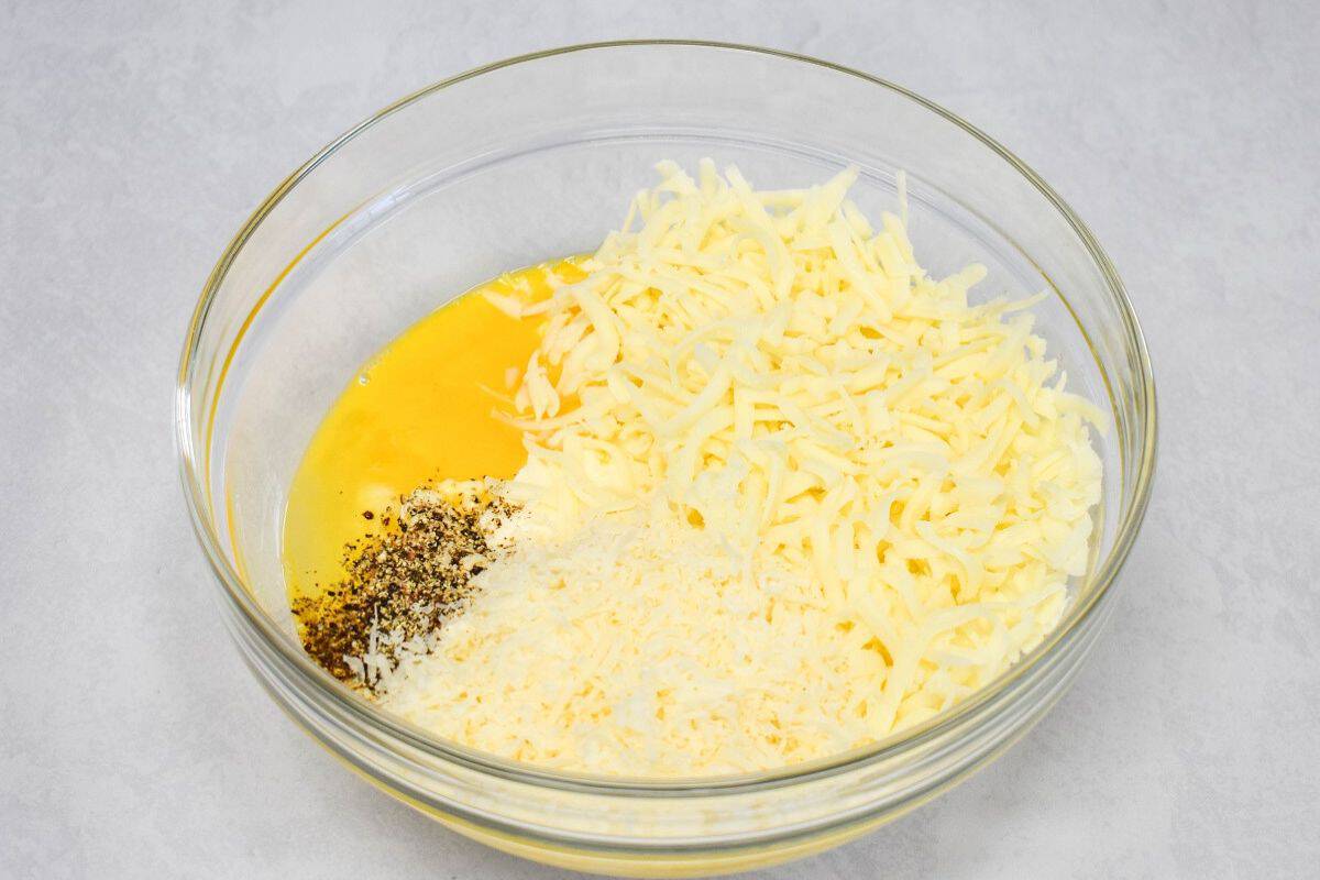 Egg, black pepper, shredded mozzarella, and grated Parmesan in a glass bowl, ready to mix with ricotta for the lasagna casserole cheese mixture.