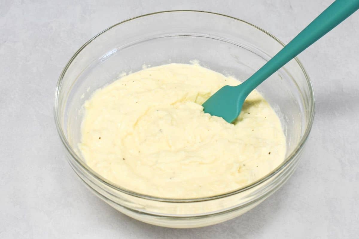 Creamy ricotta cheese mixture with black pepper in a glass bowl with a teal spatula.