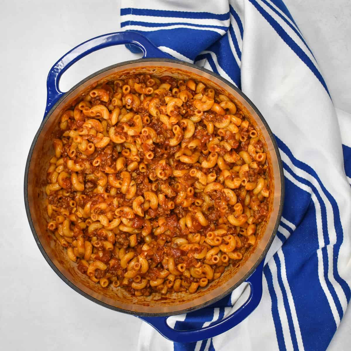 A top image of the beef and macaroni in a large blue pot set on a white table with a blue and white kitchen towel.