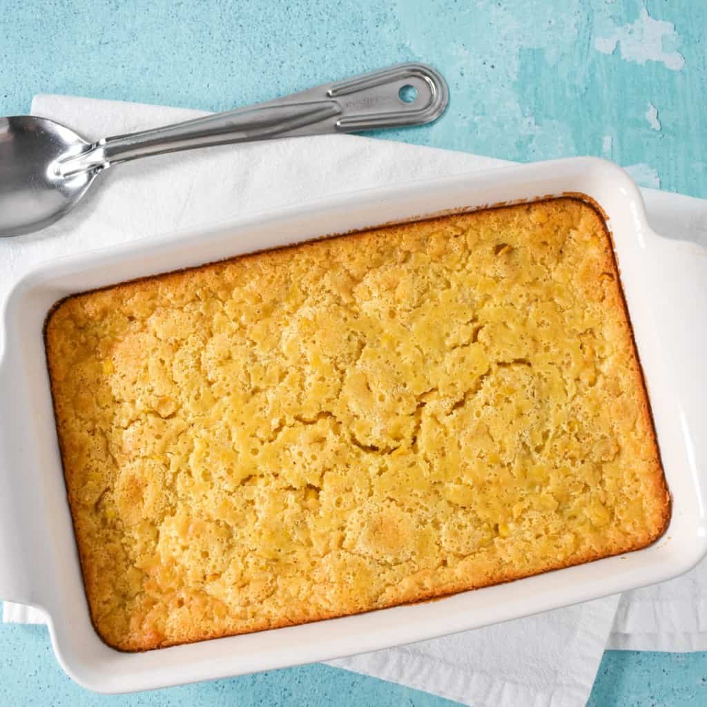 An image of the corn casserole, still in the dish and set on an aqua colored table on a white kitchen towel.