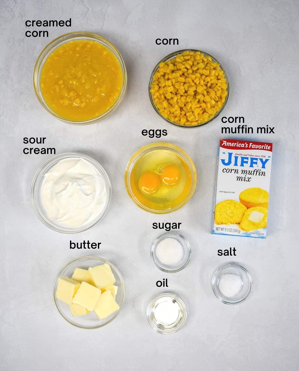 The ingredients for the casserole arranged in glass bowls on a white table, except for the corn muffin mix. Each ingredient has a  small, black label with the name above.