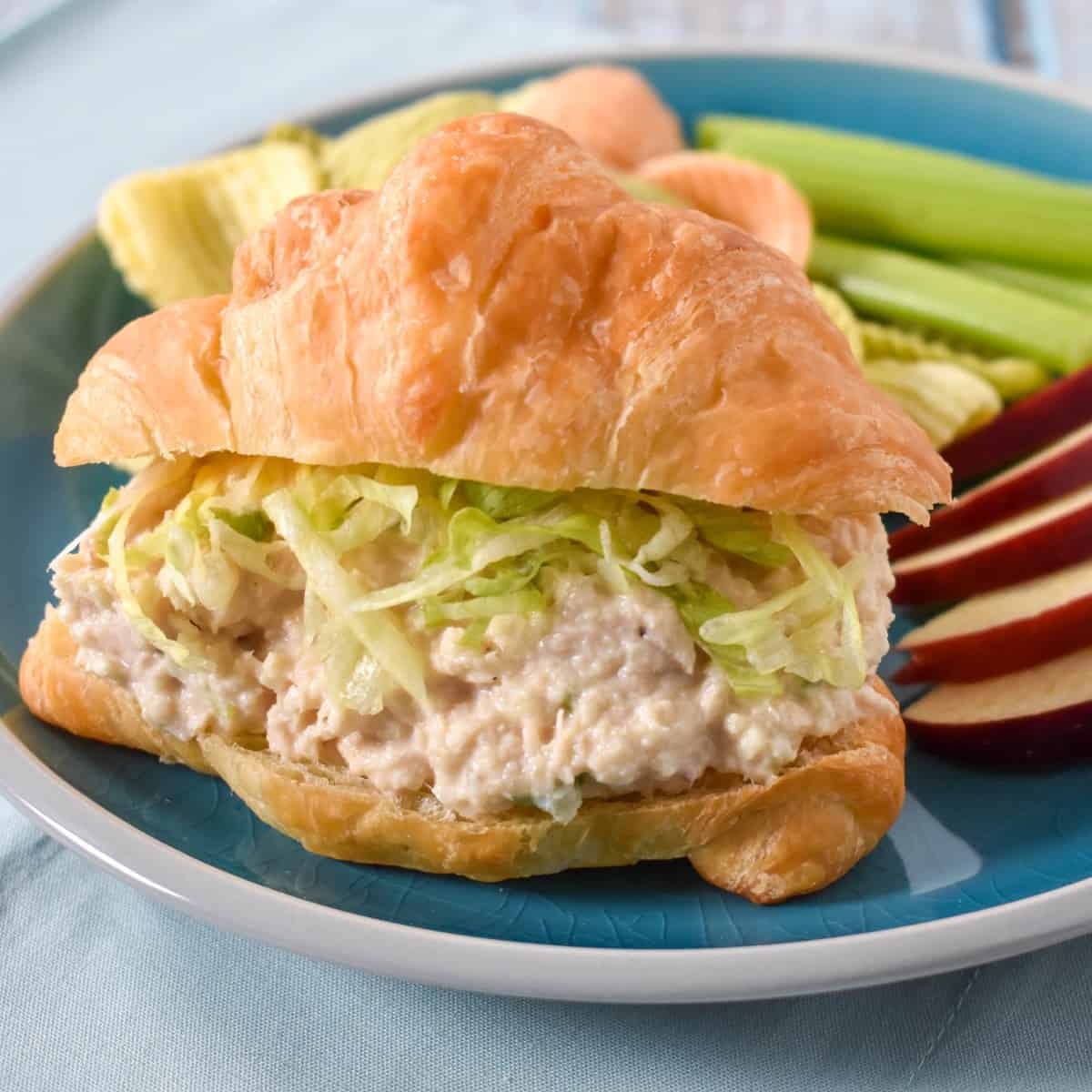 Tuna salad topped with shredded lettuce on a croissant with sliced apples and celery sticks on the side. The sandwich is served on a blue plate.