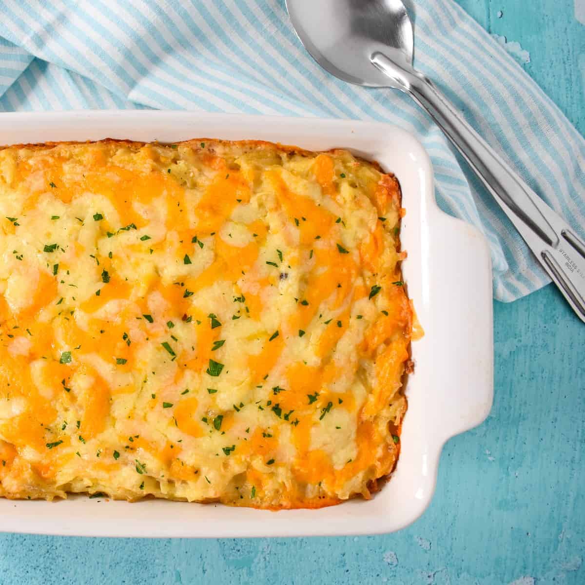 An image of the baked hash brown casserole set on a light blue table with a light blue and white striped kitchen towel and large serving spoon.