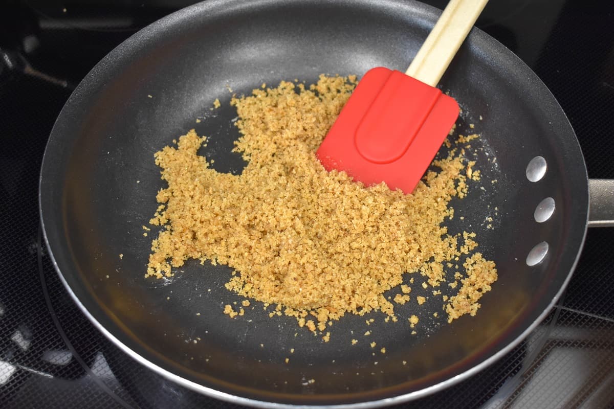 An image of breadcrumbs combined with butter in a non-stick skillet with a red silicone spatula in the skillet.