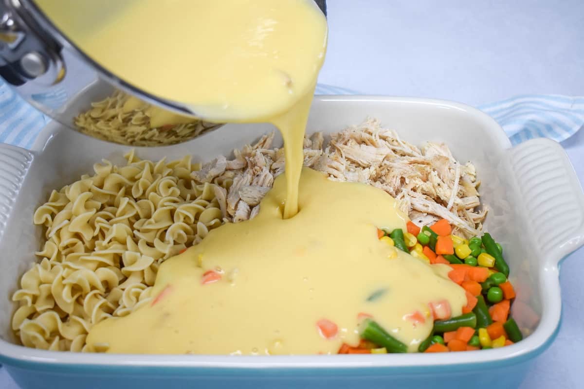 An image of the creamy sauce being added to the noodles, chicken, and vegetables in the large baking dish.