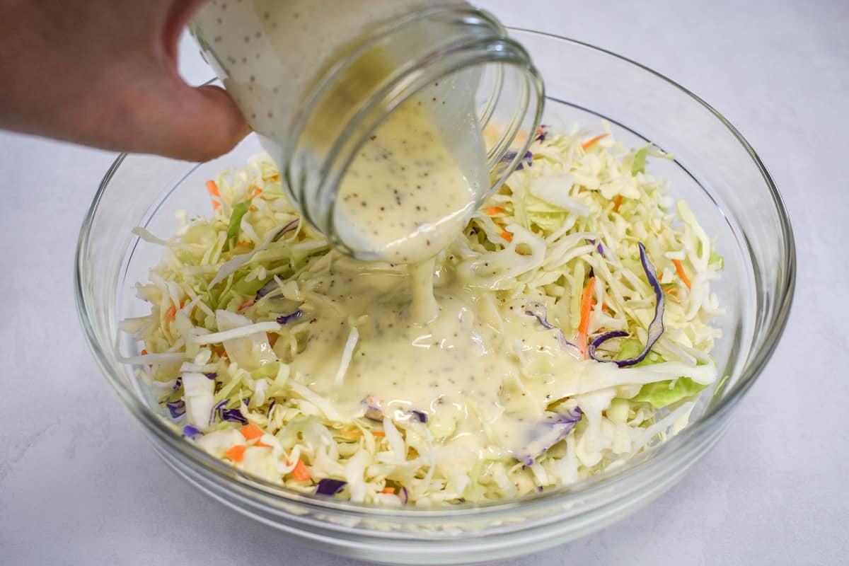 An image of dressing being added to the coleslaw mix in a large, glass bowl.