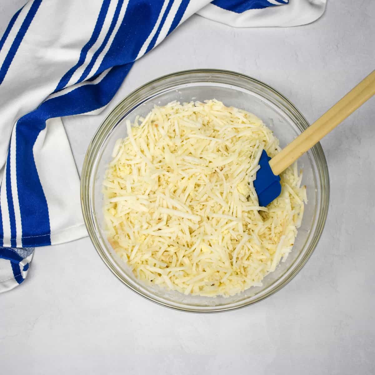 An image of the hash browns in large, glass bowl set on a white table with a blue and white striped kitchen towel.