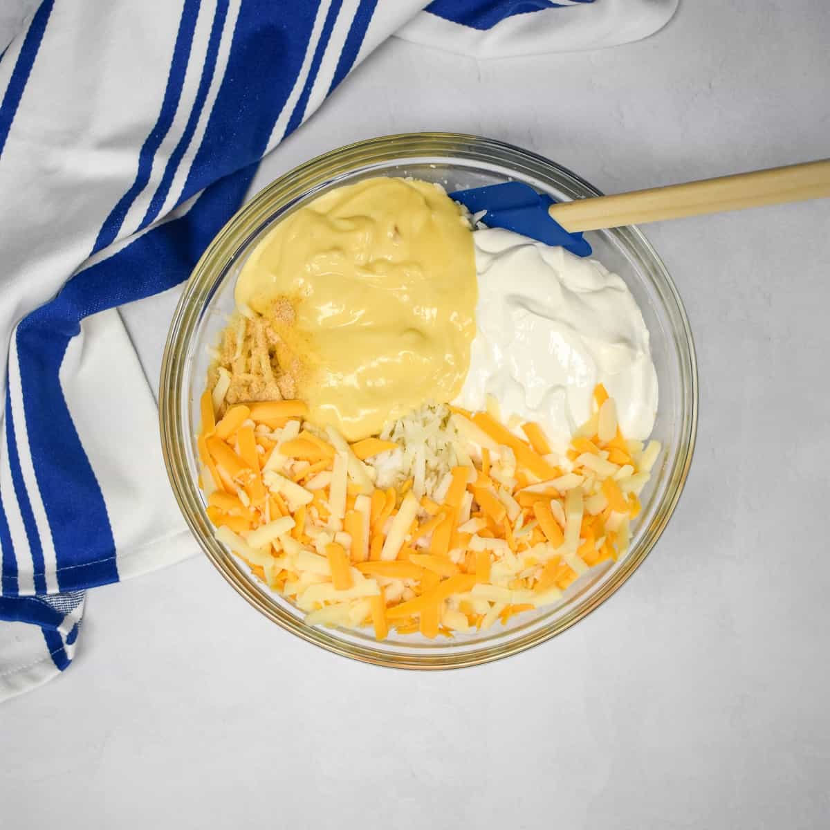 An image of the hash browns, cheese, sour cream, cream of chicken soup and seasoning in a large, glass bowl set on a white table with a blue and white striped kitchen towel.