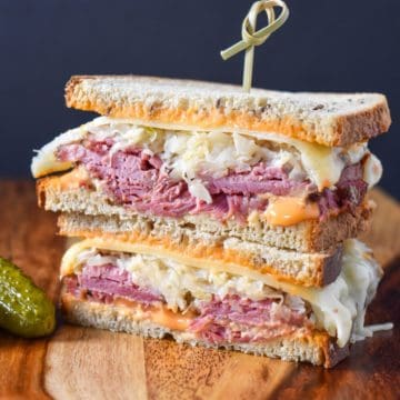 An image of a Reuben sandwich, cut in half, stacked, and held together with a toothpick. The sandwich is set on a wood cutting board.