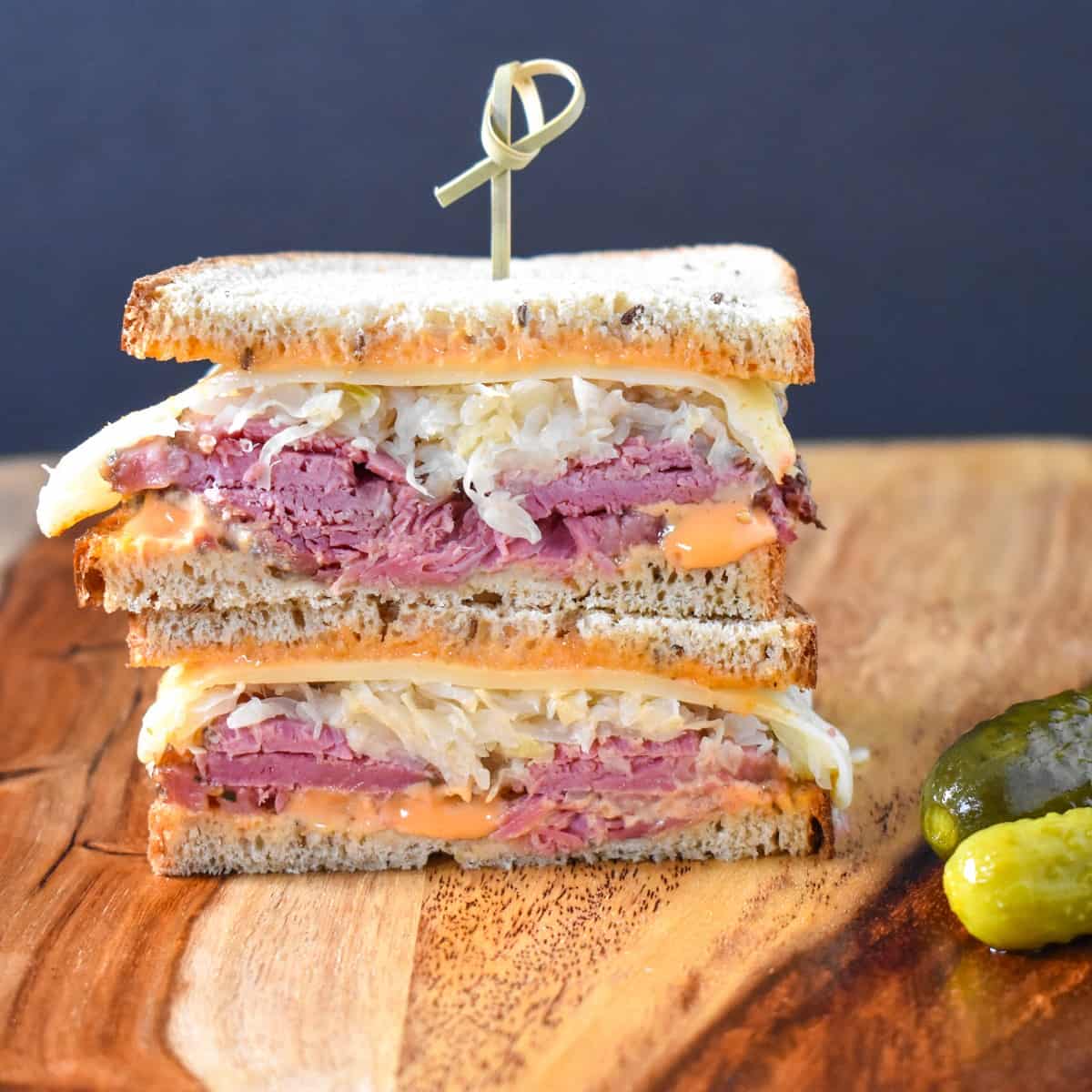 An image of a Reuben sandwich, cut in half, stacked, and held together with a toothpick. The sandwich is set on a wood cutting board.