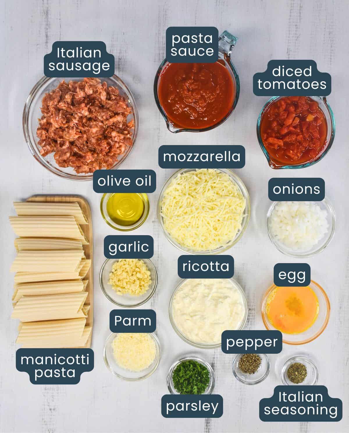 Top-down view of labeled ingredients for baked manicotti with Italian sausage, including Italian sausage, pasta sauce, diced tomatoes, mozzarella, olive oil, onions, garlic, ricotta, Parmesan, egg, pepper, parsley, Italian seasoning, and uncooked manicotti pasta.