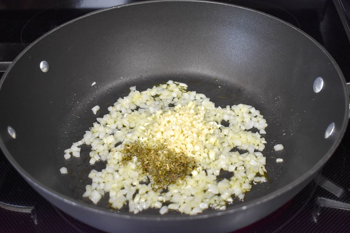An image of onions, garlic, and seasoning cooking in a large, black skillet.
