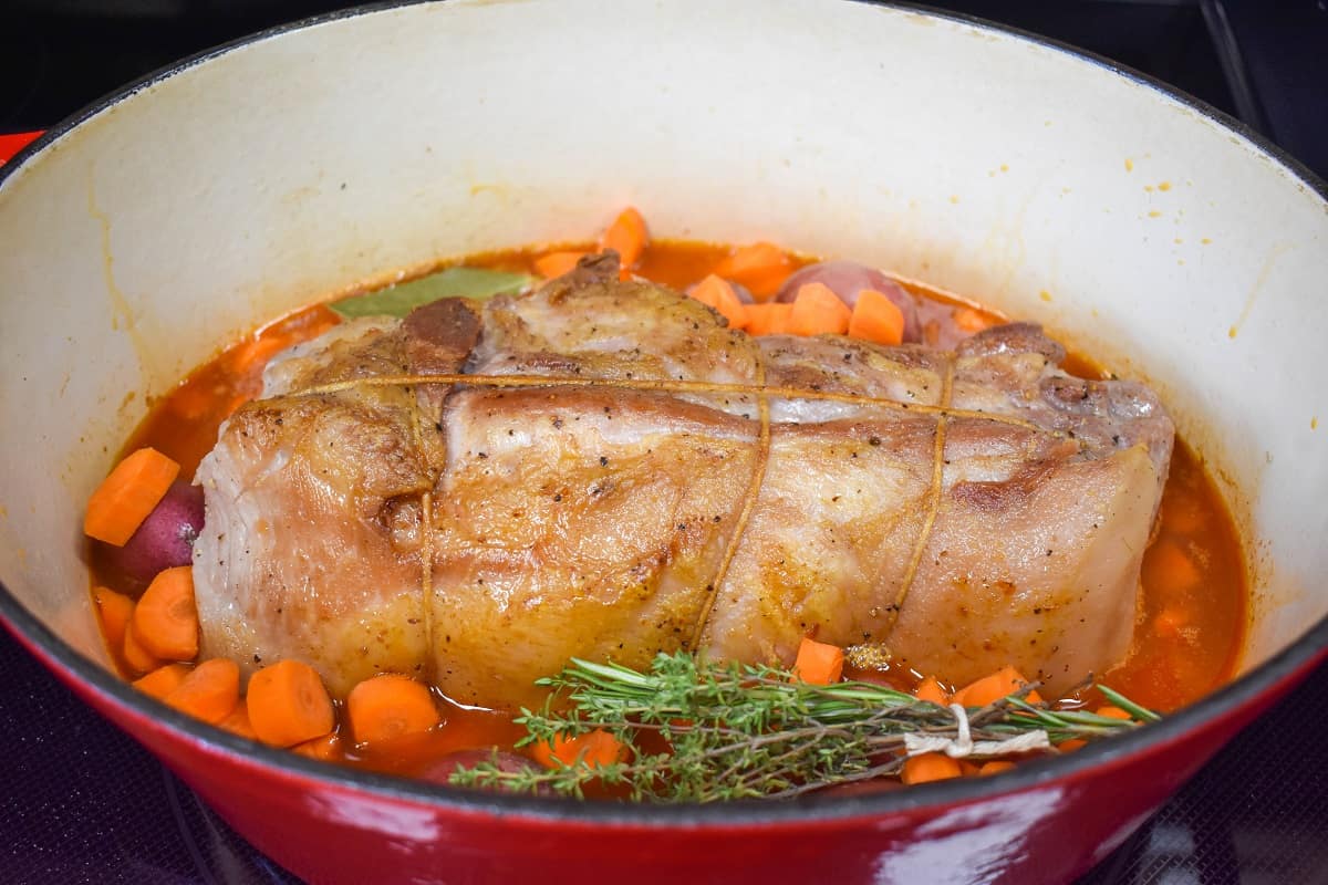 An image of the pork with the broth, sliced carrots, small red potatoes, and herbs in a large pot.