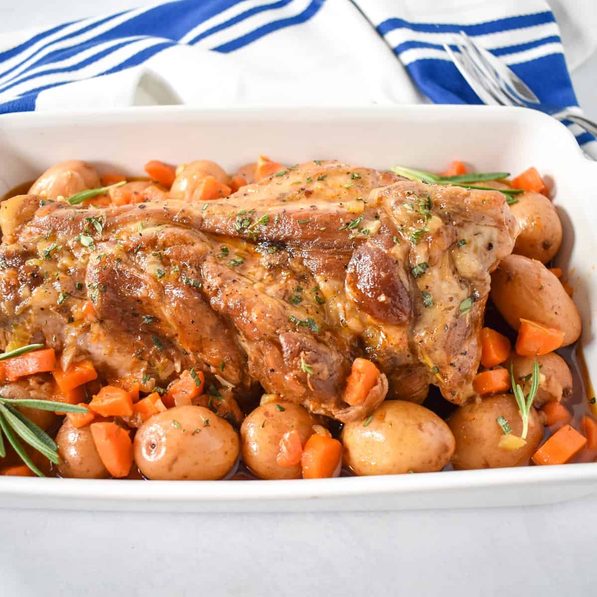 An image of the finished pork pot roast served with the potatoes and carrots in a white baking dish with a blue and white kitchen towel in the background.