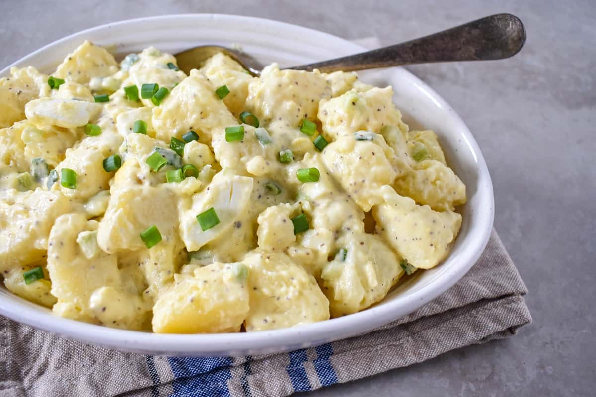 An image of the finished potato egg salad served in a white bowl with a serving spoon and set on a beige and blue linen.