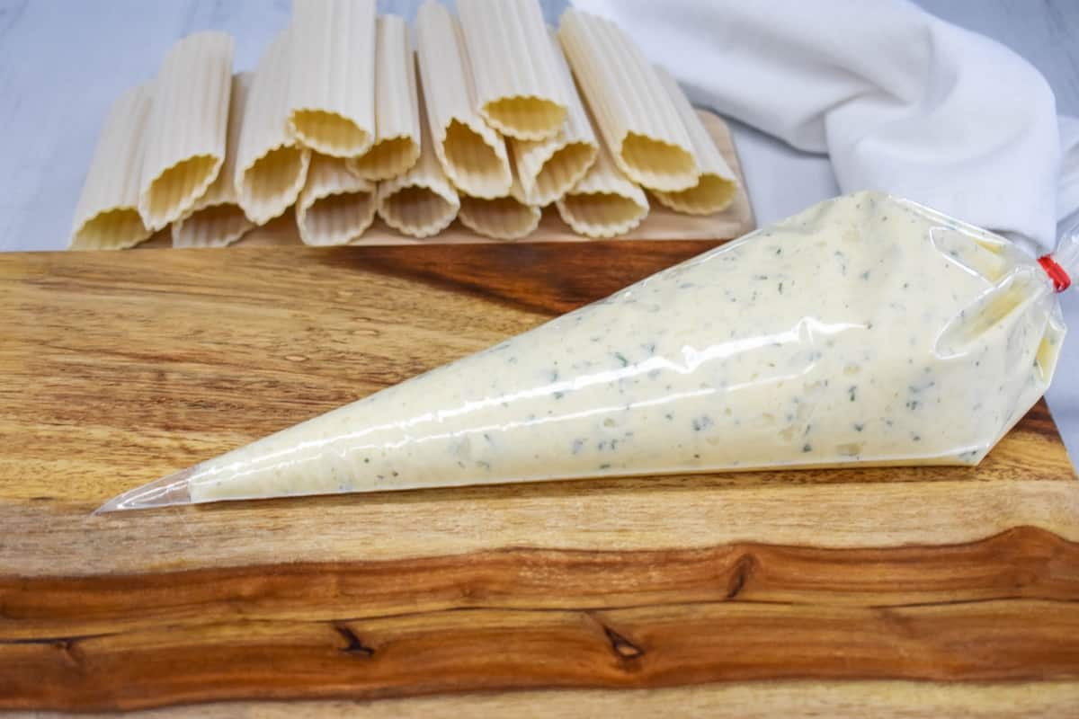 An image of a piping bag filled with the cheese mixture and set on a wood cutting board with manicotti shells in the background.
