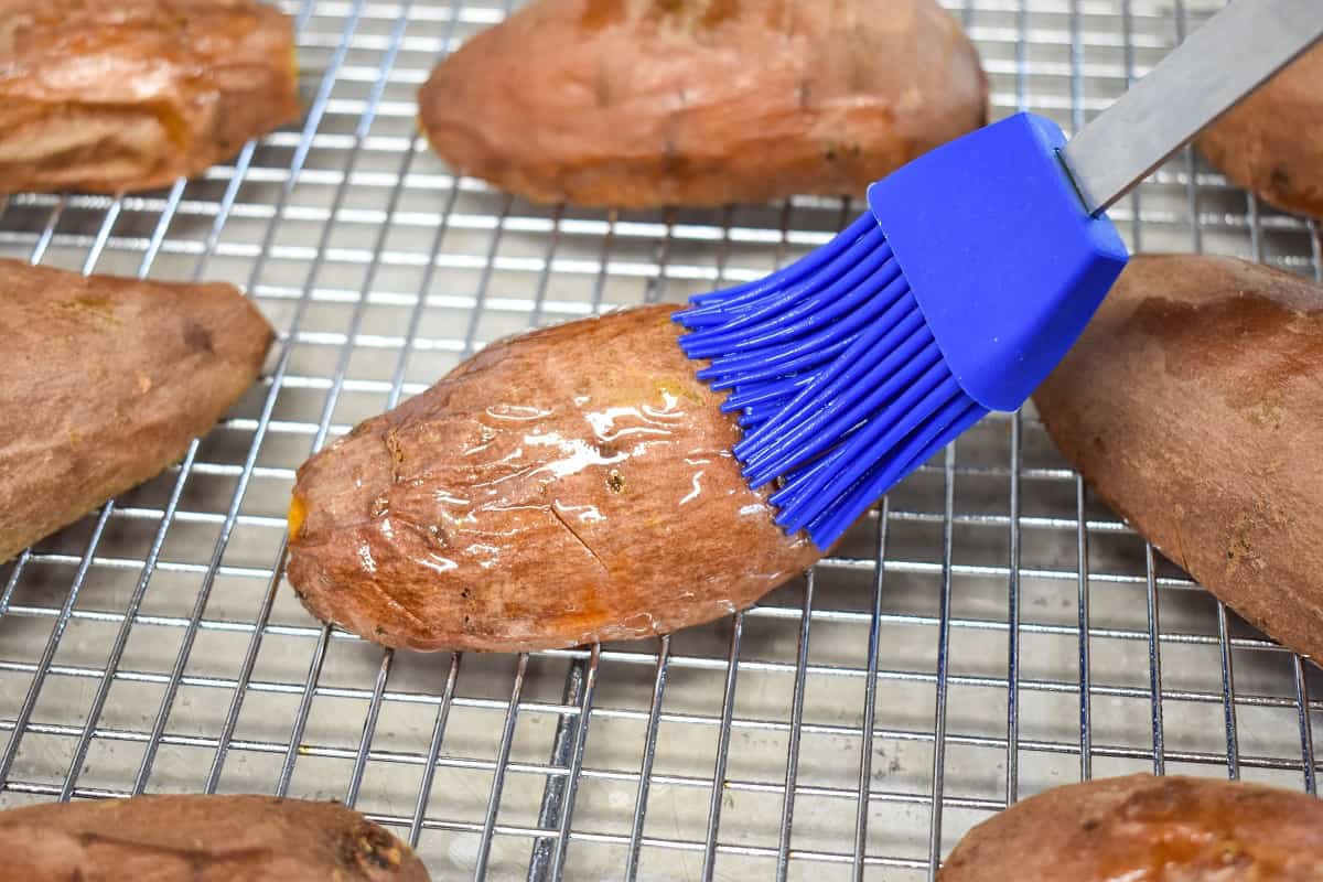 A blue silicone brush lightly coating the skin side of a halved sweet potato on a cooling rack.ng sweet potato skins image 8