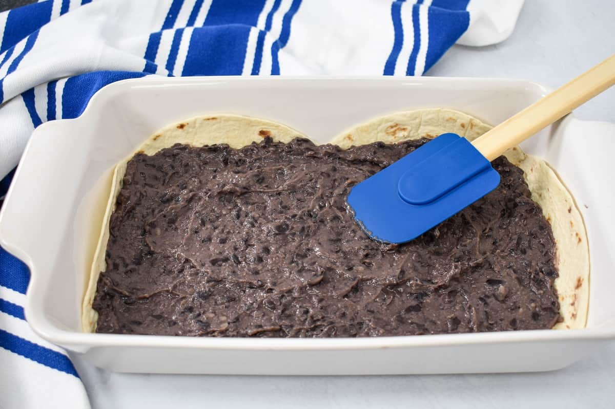Refried black beans being spread on flour tortillas in a white baking dish.