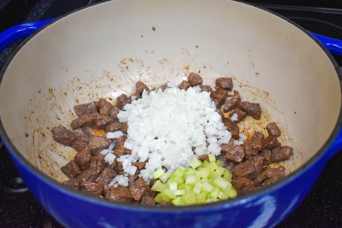 Diced onion and celery on top of browned beef pieces in a large pot.