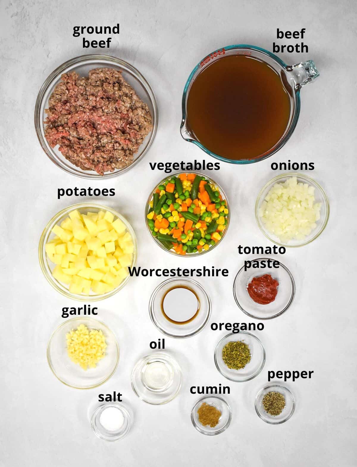 The ingredients for the soup arranged in glass bowls on a white table with each ingredient labeled in small black letters.