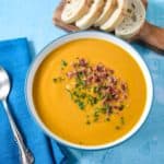 The sweet potato soup served in a white bowl with a light blue rim on a blue table with sliced bread in the background and a blue linen with a spoon to the left side.