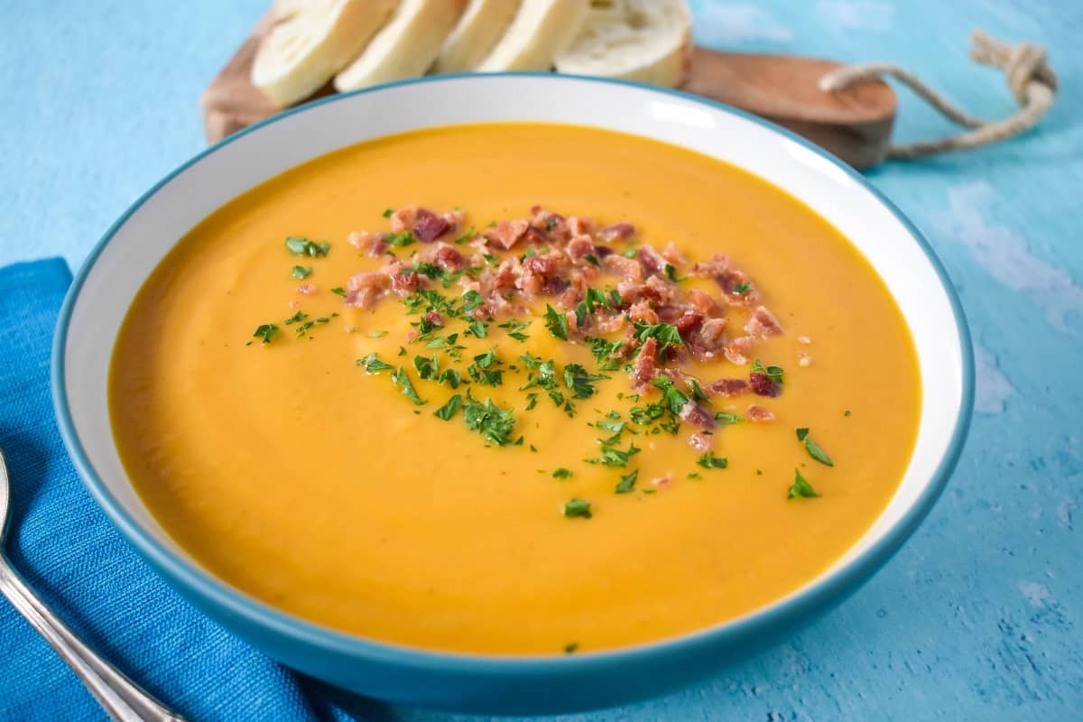 A close up of the soup garnished with crumbled bacon and parsley served in a white bowl on a light blue table with sliced bread in the background.