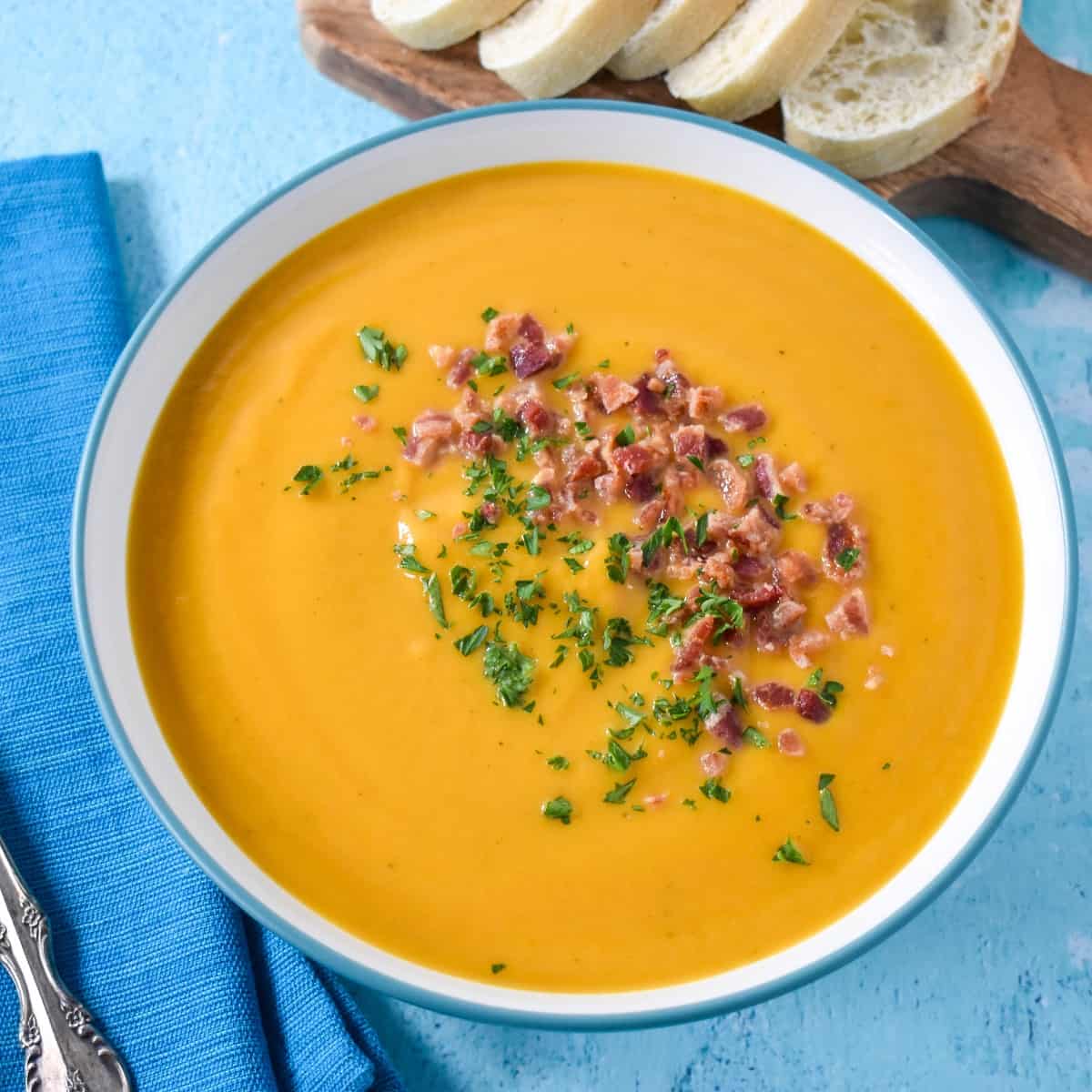 The soup garnished with crumbled bacon and parsley served in a white bowl on a light blue table with sliced bread in the background.