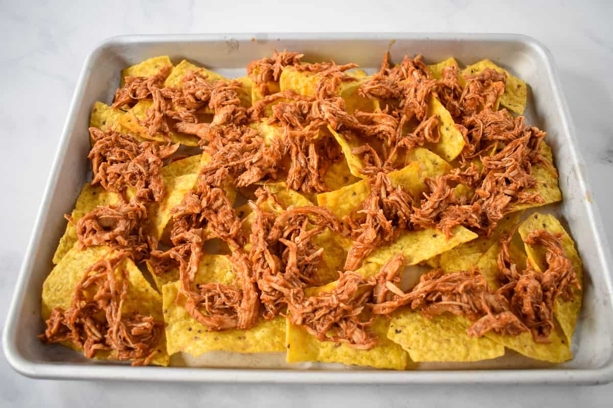 Shredded chicken arranged over the tortilla chips on a sheet pan.