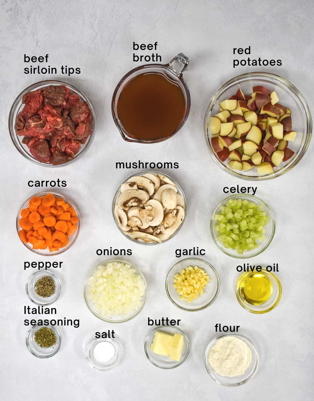 The ingredients for the stew arranged in glass bowls on a white table with each labeled with small black letters.