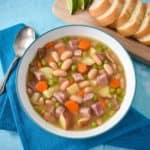 The ham and bean soup image served in a white bowl with a light blue rim set on a teal linen with sliced bread and lime wedges in the background.